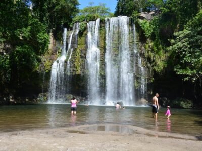 "Scenic view of Miravalles Waterfalls surrounded by lush green forest, with crystal-clear water cascading down rocky cliffs into a serene pool below."